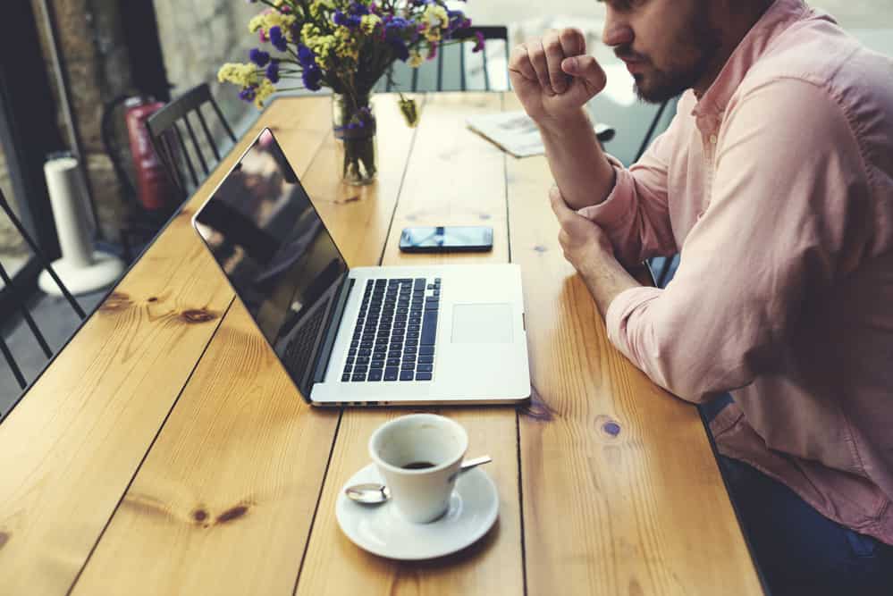 Male freelancer connecting to wireless via laptop computer, thoughtful businessman work on notebook while sitting at wooden table in modern coffee shop interior, student reading text or book in cafe